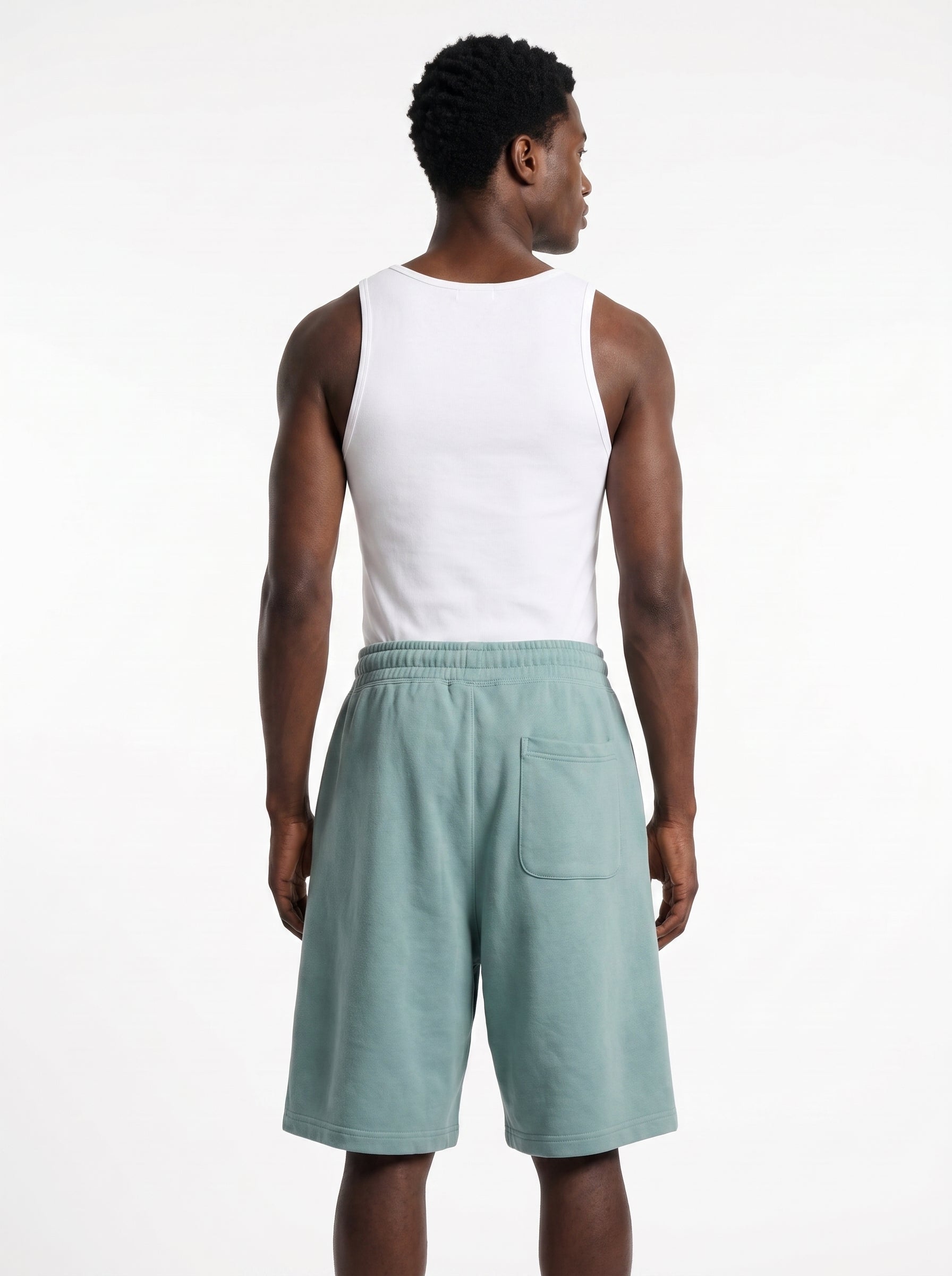 A man shown from the back wearing a white sleeveless tank top and sage green shorts against a plain white studio background.