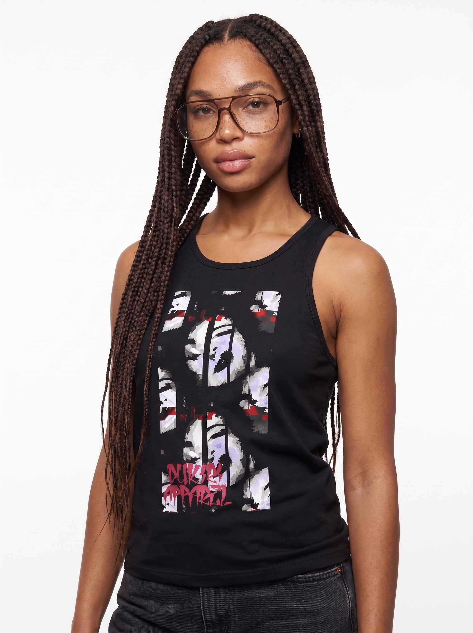 A woman with long dark braids and glasses wearing a black sleeveless tank top with a large white, black, and red graphic print on the front, paired with dark jeans, against a plain white background.