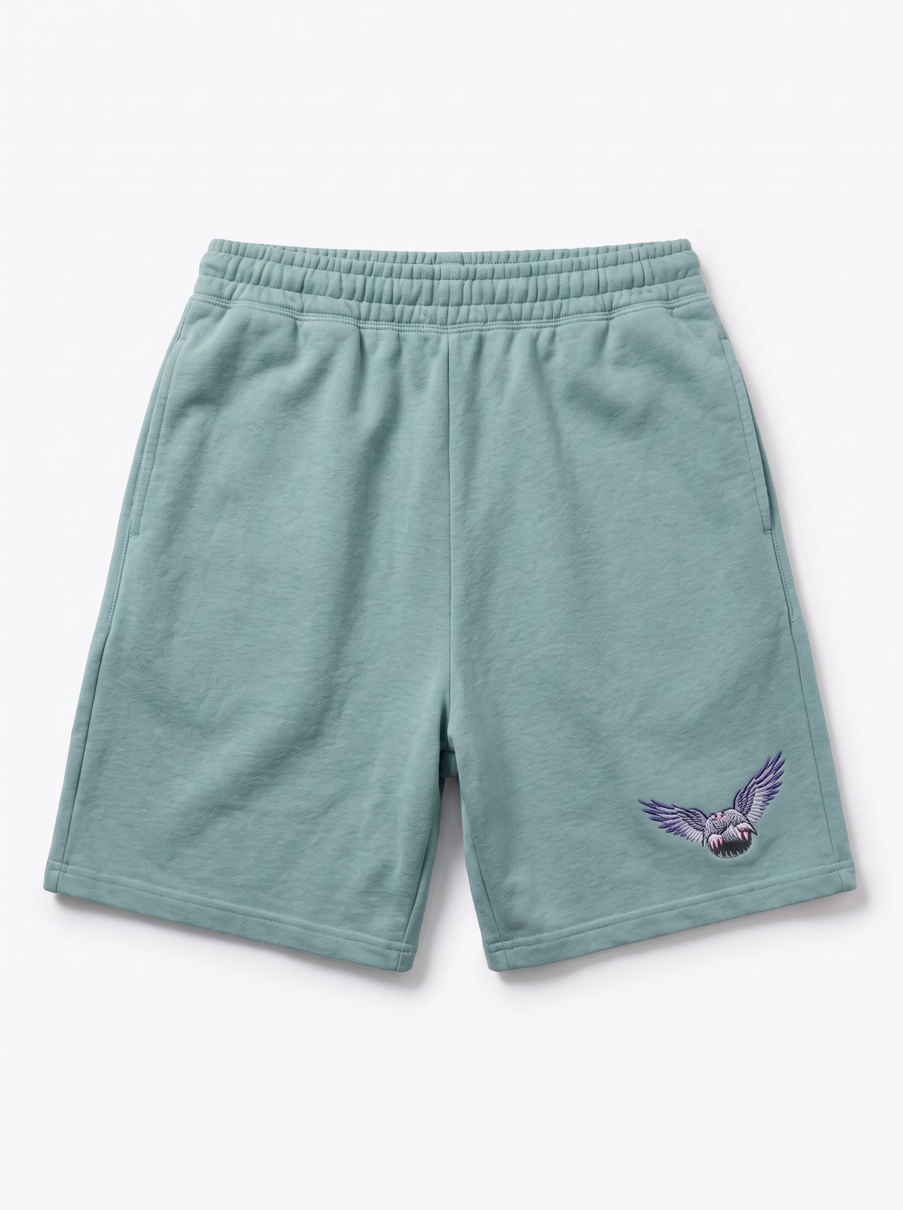 A pair of sage green sweat shorts with an embroidered purple owl graphic on the lower left leg, displayed against a clean white background.