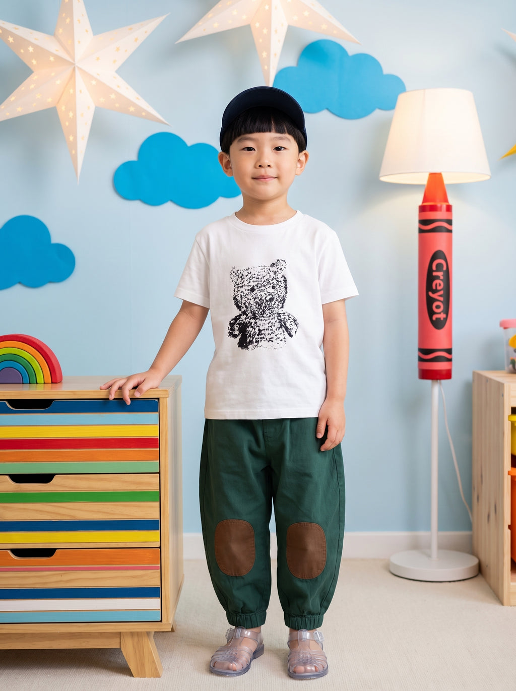 A young child wearing a white graphic t-shirt with a black bear print, dark green pants with brown knee patches, and a black cap, standing in a decorated children's room.