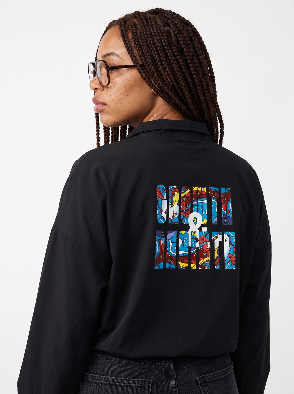 A woman with braided hair and glasses, shown from the back, wearing a black long-sleeve shirt with a colorful graphic print on the back. The print features the text 'GROWTH & REBIRTH' in a multicolor abstract block pattern.