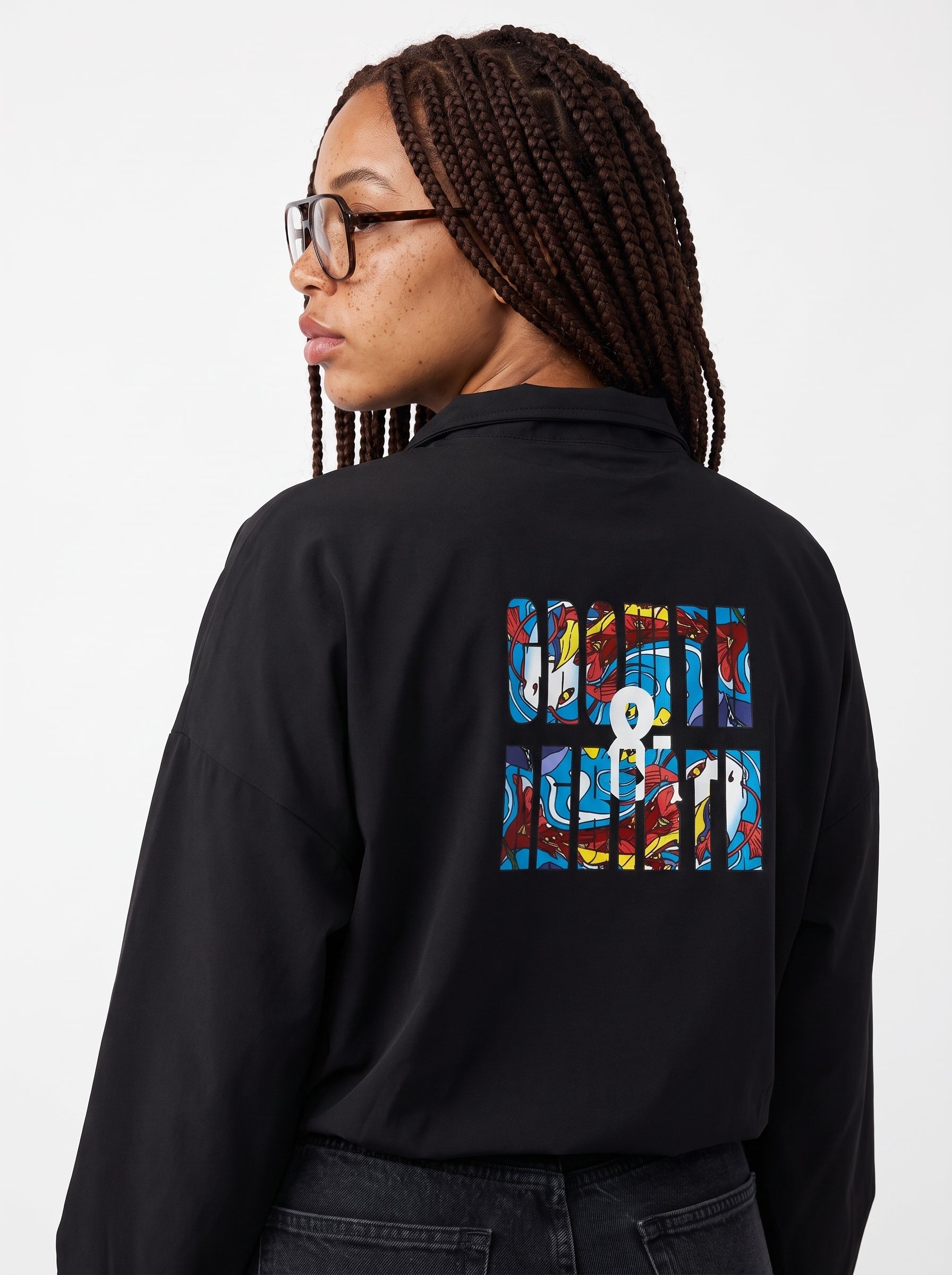 A woman with braided hair and glasses, shown from the back, wearing a black long-sleeve shirt with a colorful graphic print on the back. The print features the text 'GROWTH & REBIRTH' in a multicolor abstract block pattern.
