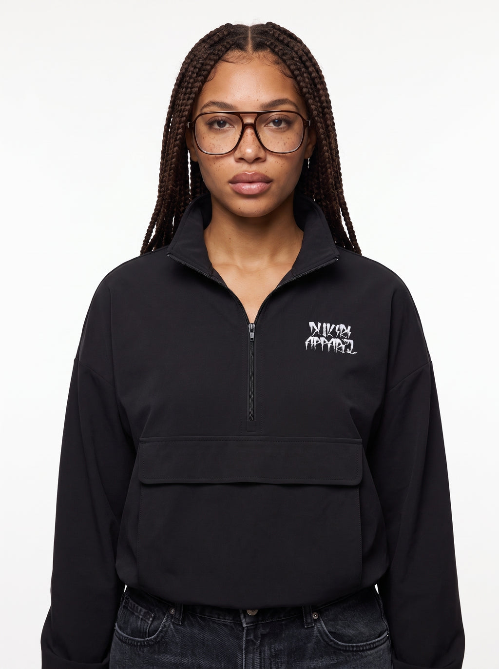A woman with braided hair and glasses wearing a black half-zip pullover sweatshirt with a white Dukiri Apparel graphic logo on the left chest, paired with dark jeans, against a plain white background.