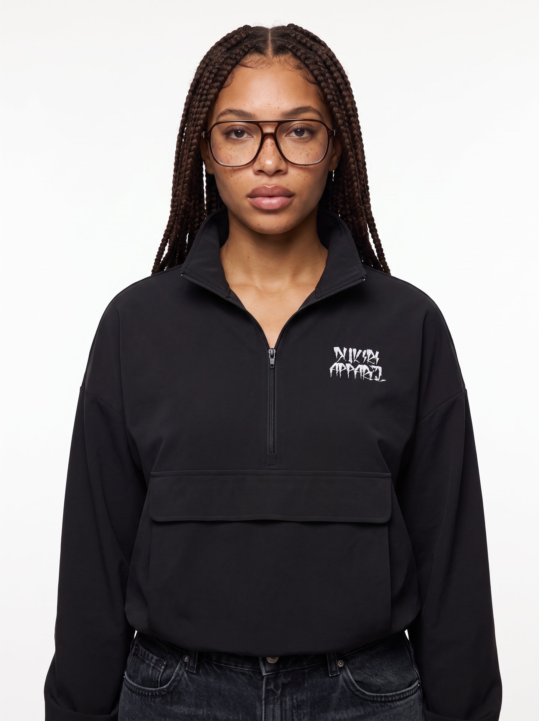 A woman with braided hair and glasses wearing a black half-zip pullover sweatshirt with a white Dukiri Apparel graphic logo on the left chest, paired with dark jeans, against a plain white background.