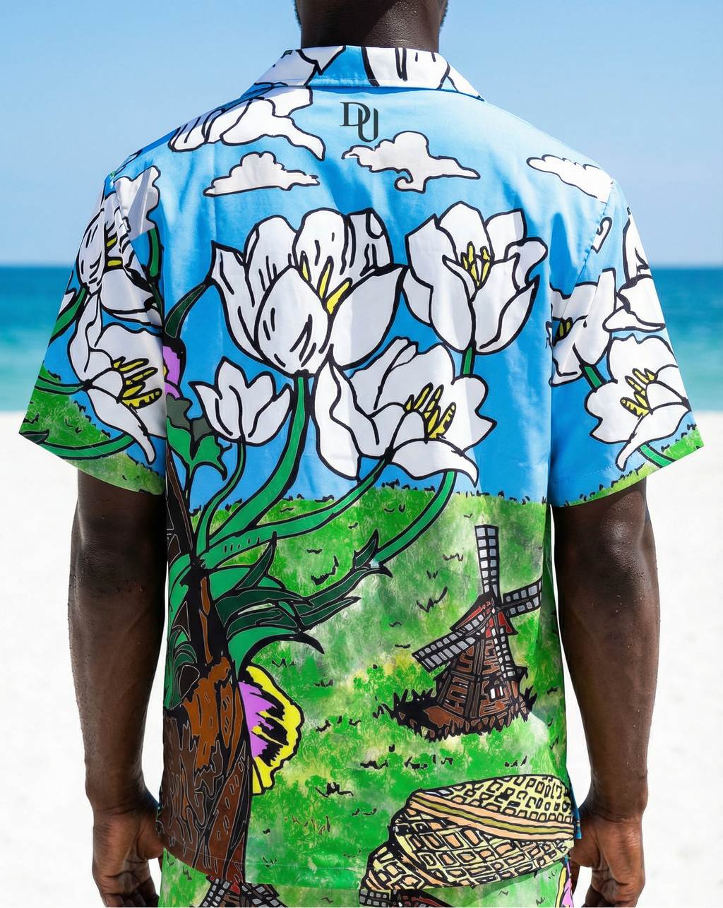 Person wearing a colorful shirt with floral and windmill design on a beach.