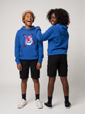 Two children modeling bright blue hoodies in a studio setting. The child on the left wears a tan beanie, black cargo shorts, dark socks, and white sneakers, while the child on the right wears black cargo shorts and black sneakers.