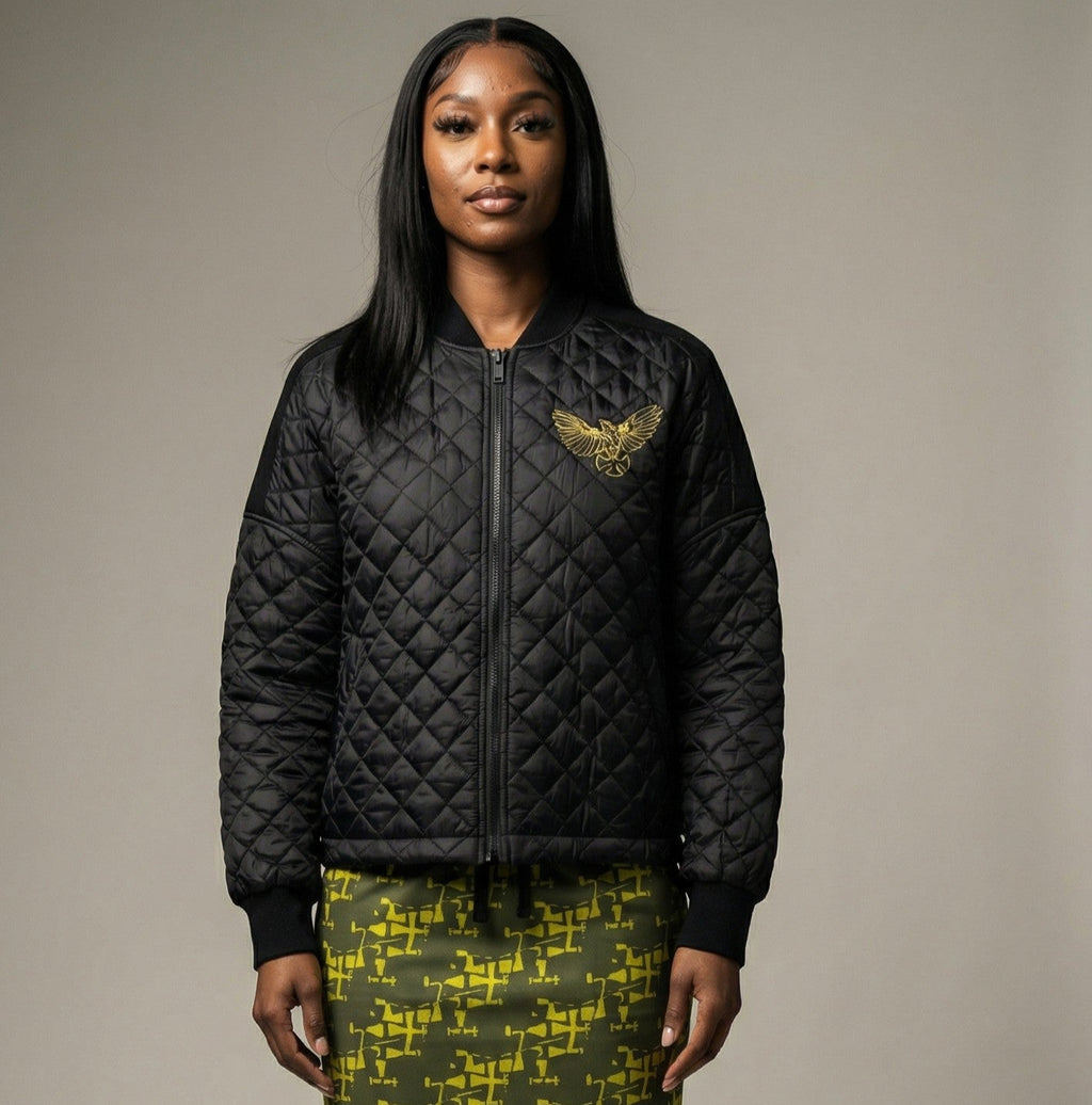 Woman wearing a black quilted jacket with a gold eagle emblem on a neutral background